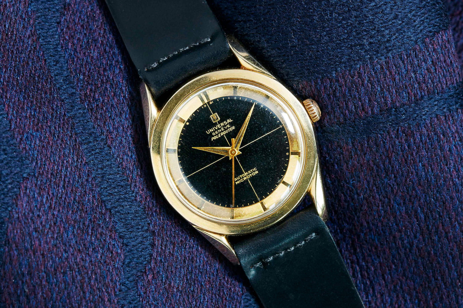 The Universal Genève Polerouter Gold Plate by Universal Geneve features a gold case, black dial with gold markers and hands, and a black leather strap, shown resting elegantly on textured dark blue and purple fabric.