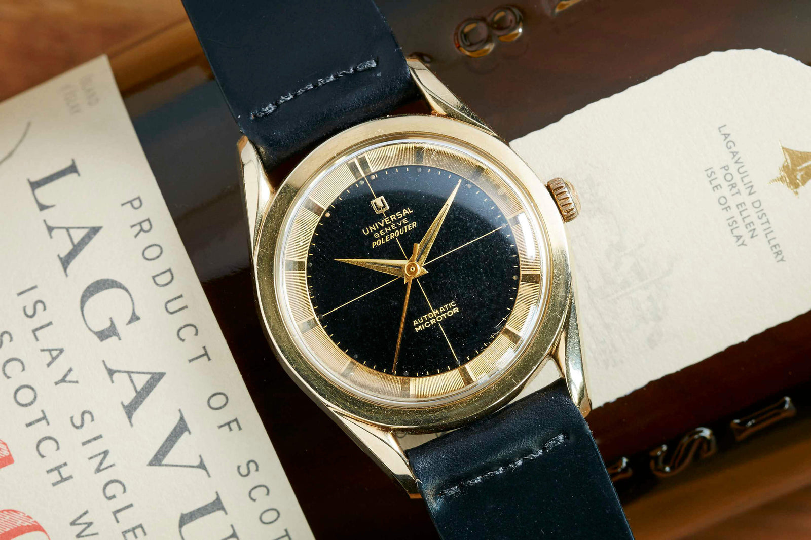 A Universal Geneve Polerouter Gold Plate vintage dress watch with a black leather strap rests on a Lagavulin Scotch whisky bottle, showcasing its black and gold face and the bottle's embossed label.