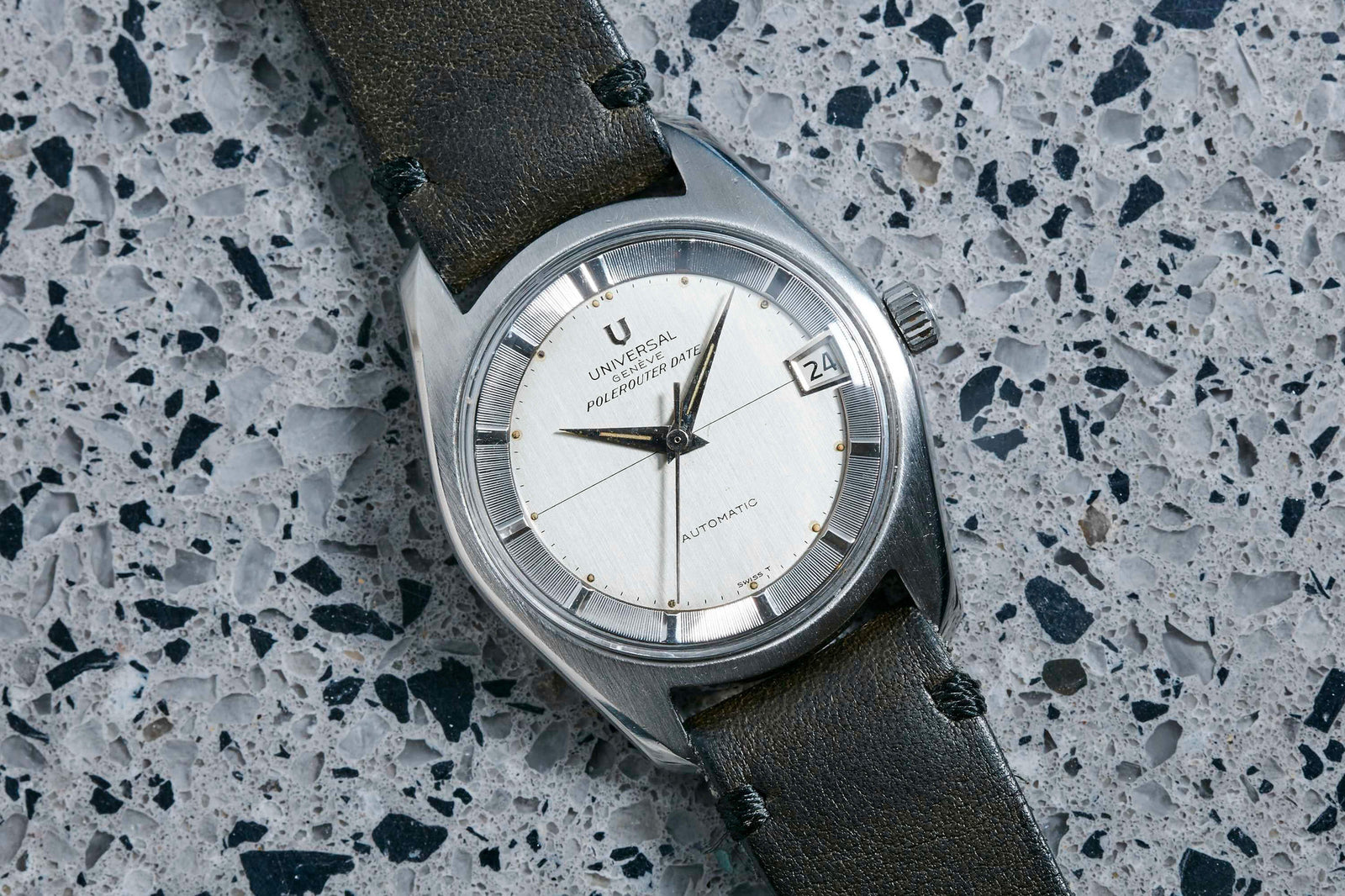 A Universal Geneve Polerouter wristwatch with a silver dial, black leather strap, and date window sits on a speckled gray and black stone surface. Designed by Gerald Genta.