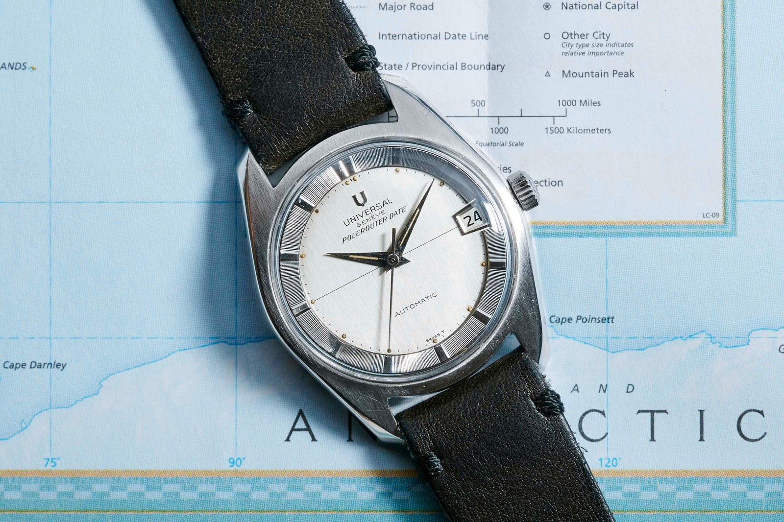 A Universal Geneve Polerouter wristwatch with a black leather strap rests on a map of Antarctica. Designed by Gerald Genta, it has a silver-white dial, black markers, and a date window at 3 o'clock.