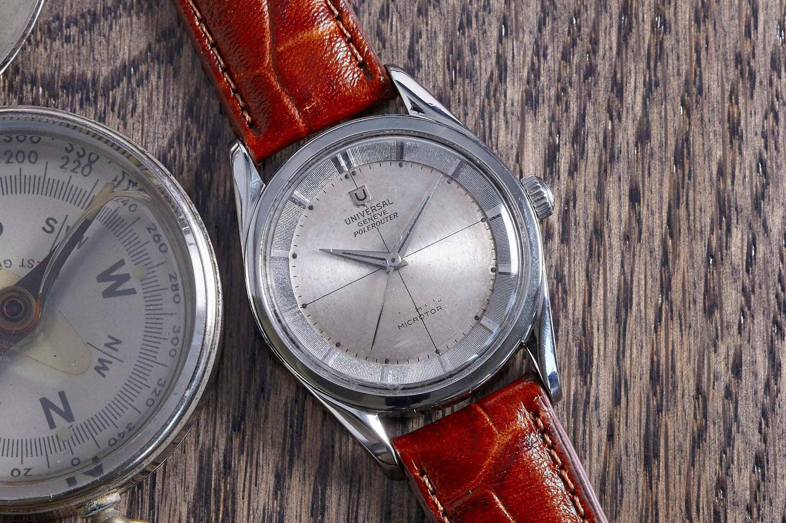 A Universal Geneve Polerouter with a silver dial and brown leather strap rests on wood beside a metal compass, showcasing the timeless style of this iconic luxury stainless steel sports watch.