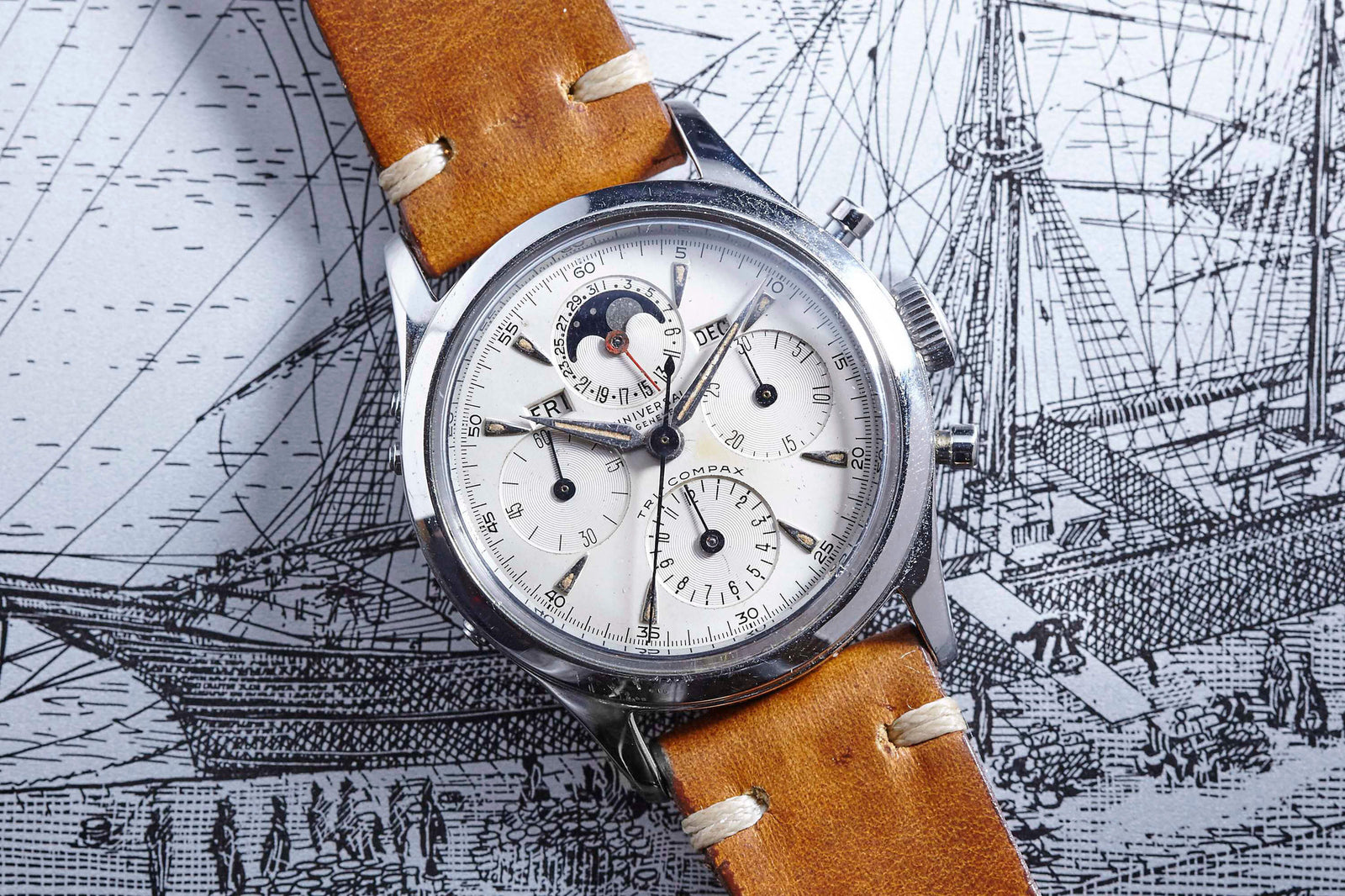 A Universal Genève Tri-Compax Moonphase wristwatch with a brown leather strap and multiple dials, including a moonphase feature, is shown resting on an intricate black-and-white illustration of a sailing ship.
