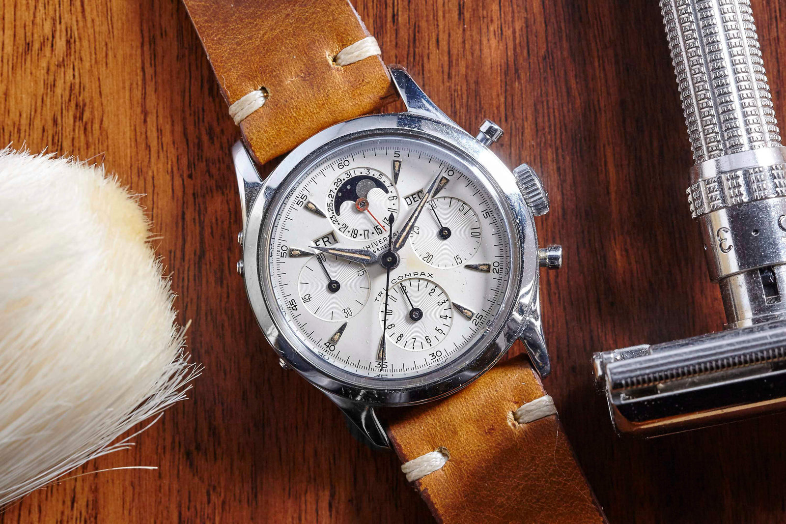 A Universal Geneve Tri-Compax Moonphase wristwatch with a tan leather strap sits on wood, alongside a shaving brush and metal safety razor. The vintage watch displays multiple dials and a distinctive moonphase complication.