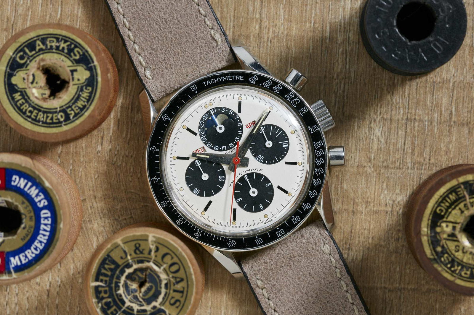 A Universal Geneve Tri-Compax chronograph wristwatch with a brown leather strap rests on a wooden surface, surrounded by vintage sewing thread spools with old-fashioned labels.
