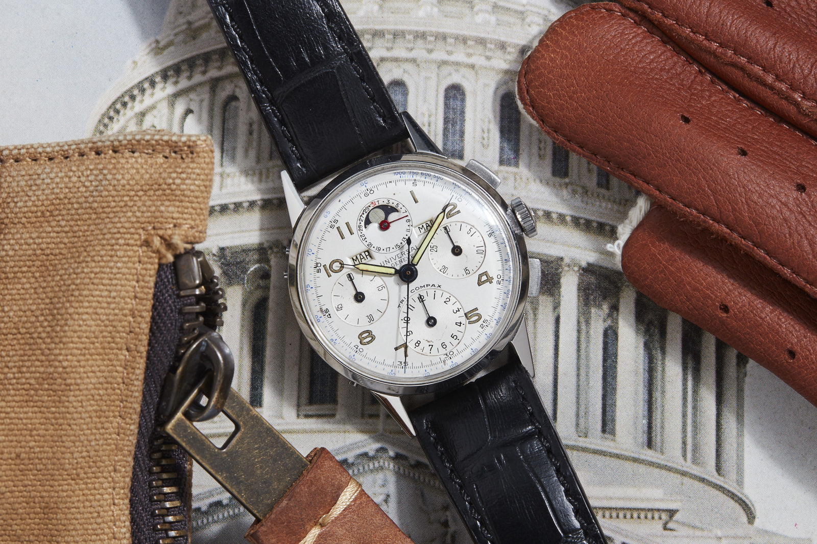 A Universal Geneve Tri-Compax luxury watch with a white dial and black leather strap is shown next to a glove and beige pouch, set against a US Capitol photo—ideal as an elegant gift.