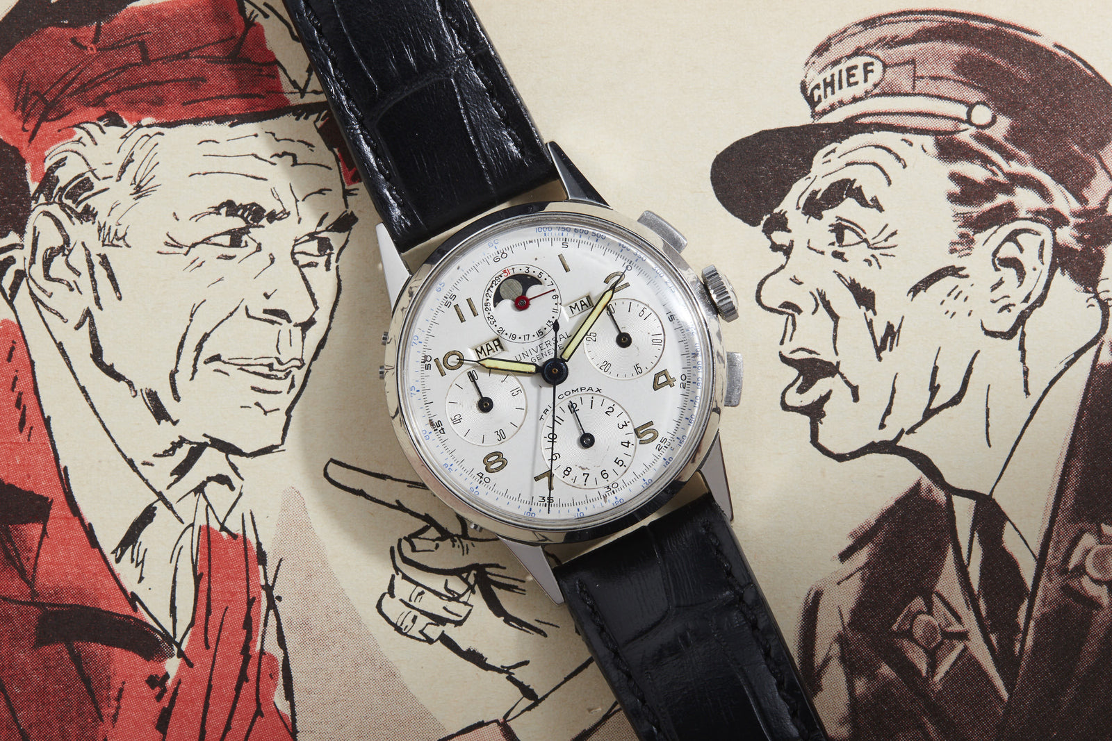 A Universal Geneve Tri-Compax vintage wristwatch with a black leather strap rests on a comic-style illustration of two men, one in a hat and coat, the other wearing a CHIEF cap.