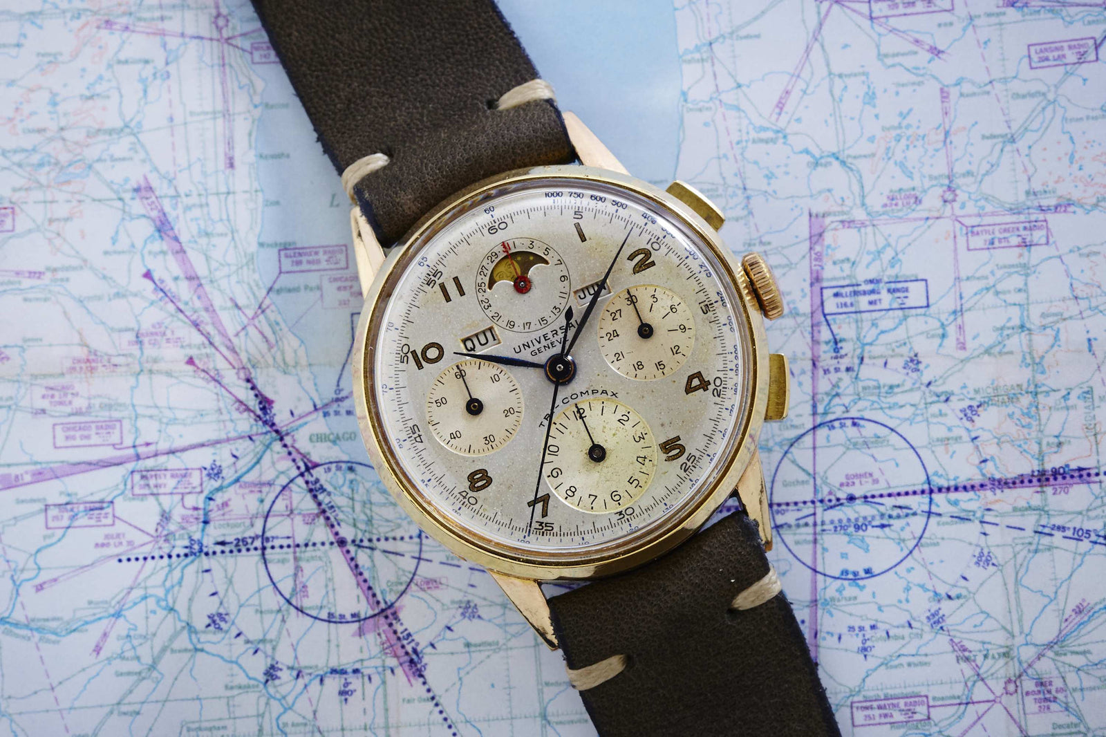 The Universal Genève Tricompax - 42406, a vintage watch with a brown leather strap and multi-dial face featuring moon phase and calendar, sits atop an aviation map marked in blue. Detailed description coming soon!.
