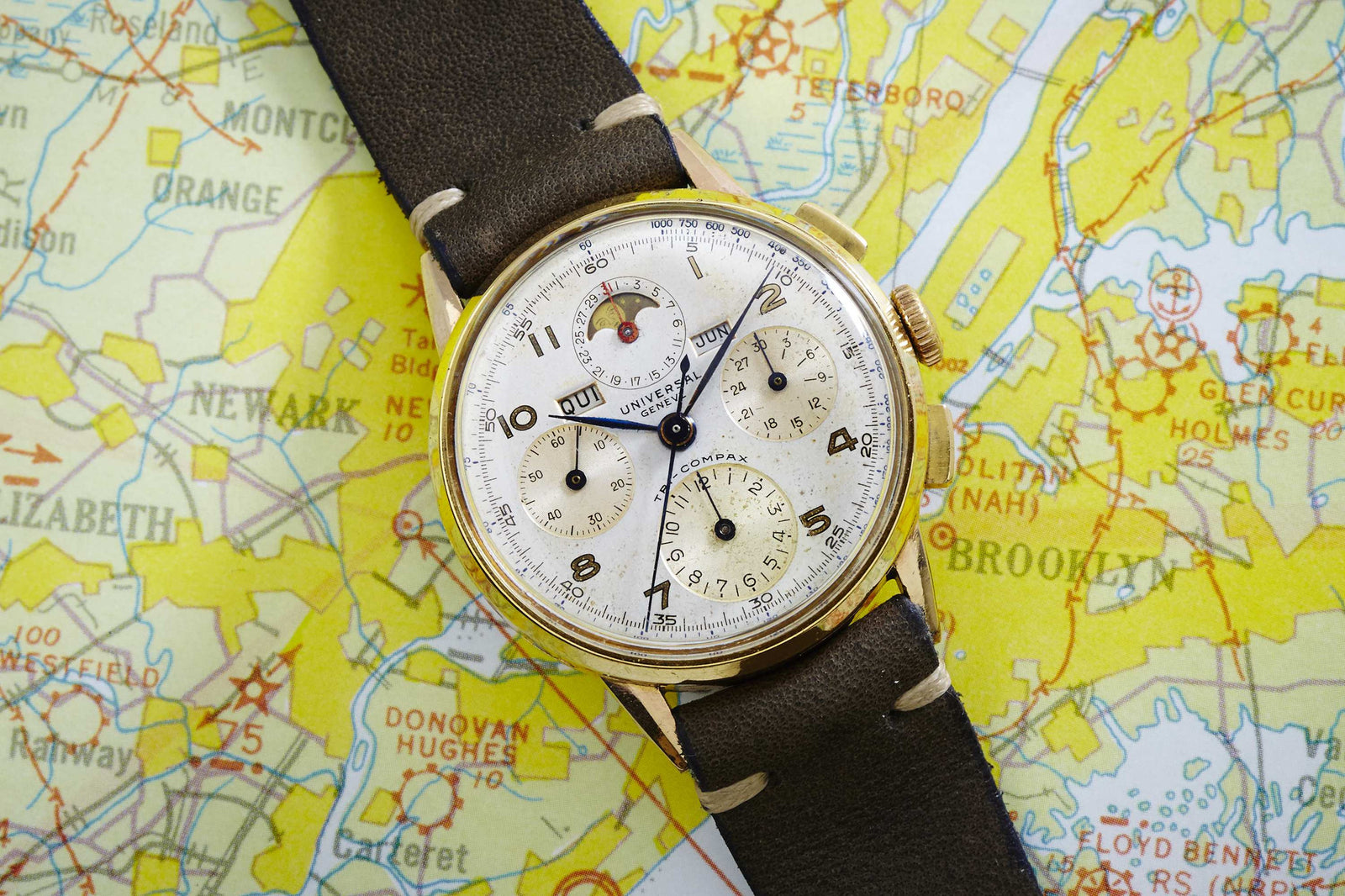 The Universal Genève Tricompax - 42406 by Universal Geneve features a gold-tone case and white dial with detailed subdials and calendar, complemented by a brown leather strap. Shown resting on a vibrant aviation map. Detailed Description Coming Soon!.