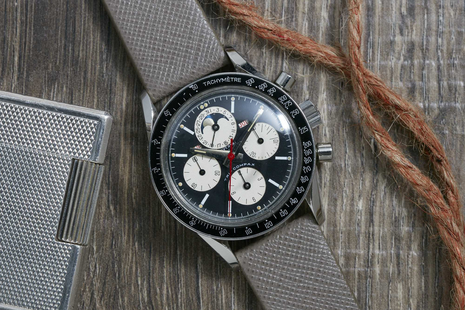 The Universal Genève Tri-Compax wristwatch features a gray textured strap, black bezel, and black dial with three white subdials and a tachymeter scale, displayed on wood beside metal and rope, echoing classic Universal Geneve chronographs.