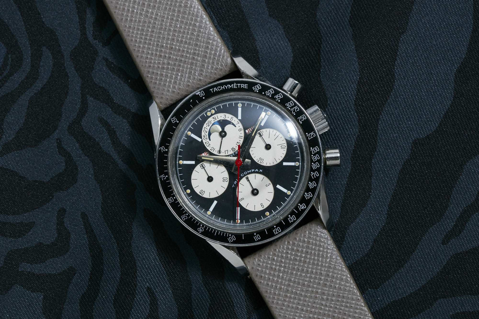 The Universal Genève Tri-Compax by Universal Geneve is a luxury chronograph wristwatch with a black and white dial, three subdials, moonphase display, tachymeter bezel, and beige textured strap, set against a dark patterned background.
